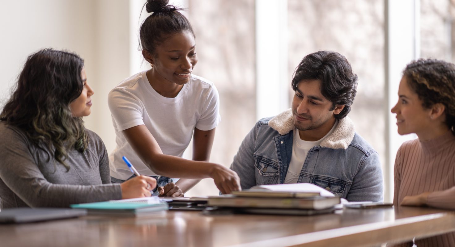 A young female University student of African decent, stands behind a peer and leans in as she tries to help her classmate with his studies. They are both dressed casually and are among a group sitting at the table all studying together for class.